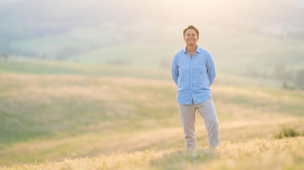 Une personne souriante debout dans un champ de hautes herbes sous une douce lumière dorée au coucher du soleil.